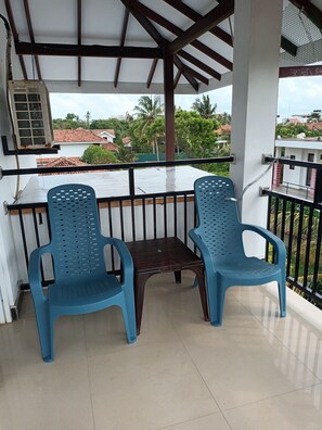 A covered balcony with two blue plastic chairs and a small table, overlooking a residential area with red-tiled roofs and greenery.