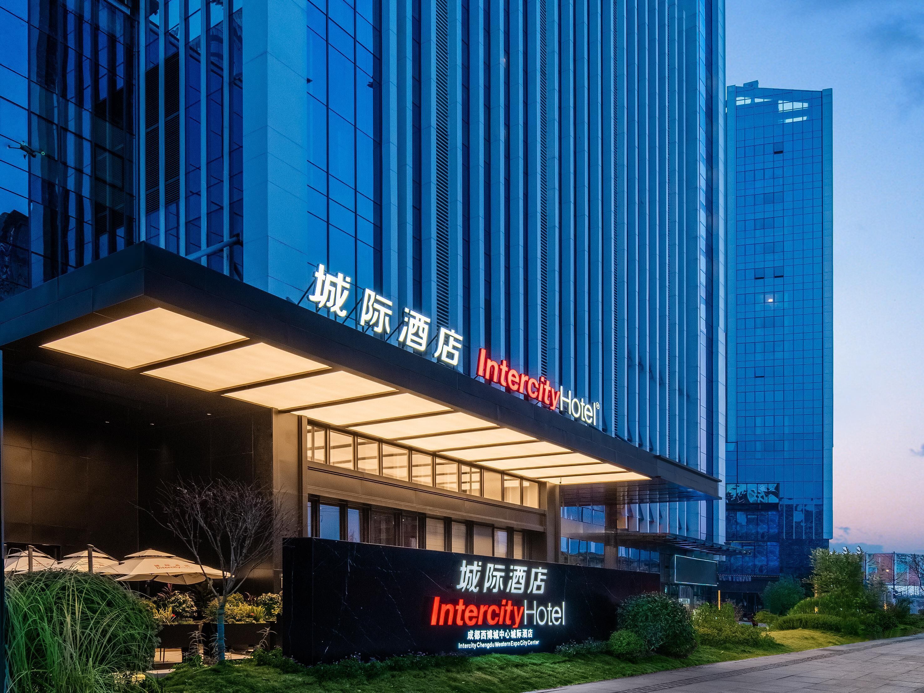 A modern hotel building with illuminated signage, large windows, and a well-maintained outdoor area with greenery.