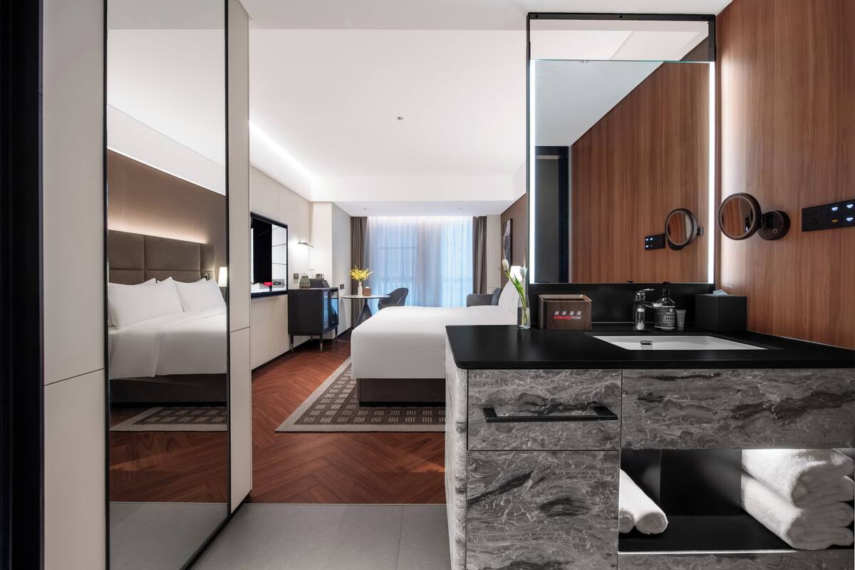 Una habitación de hotel moderna con una cama grande, una zona de estar separada, un baño con tocador y un espejo grande.