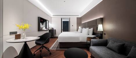 A modern hotel room with a bed, a desk, a chair, a sofa, and a small table with a vase.