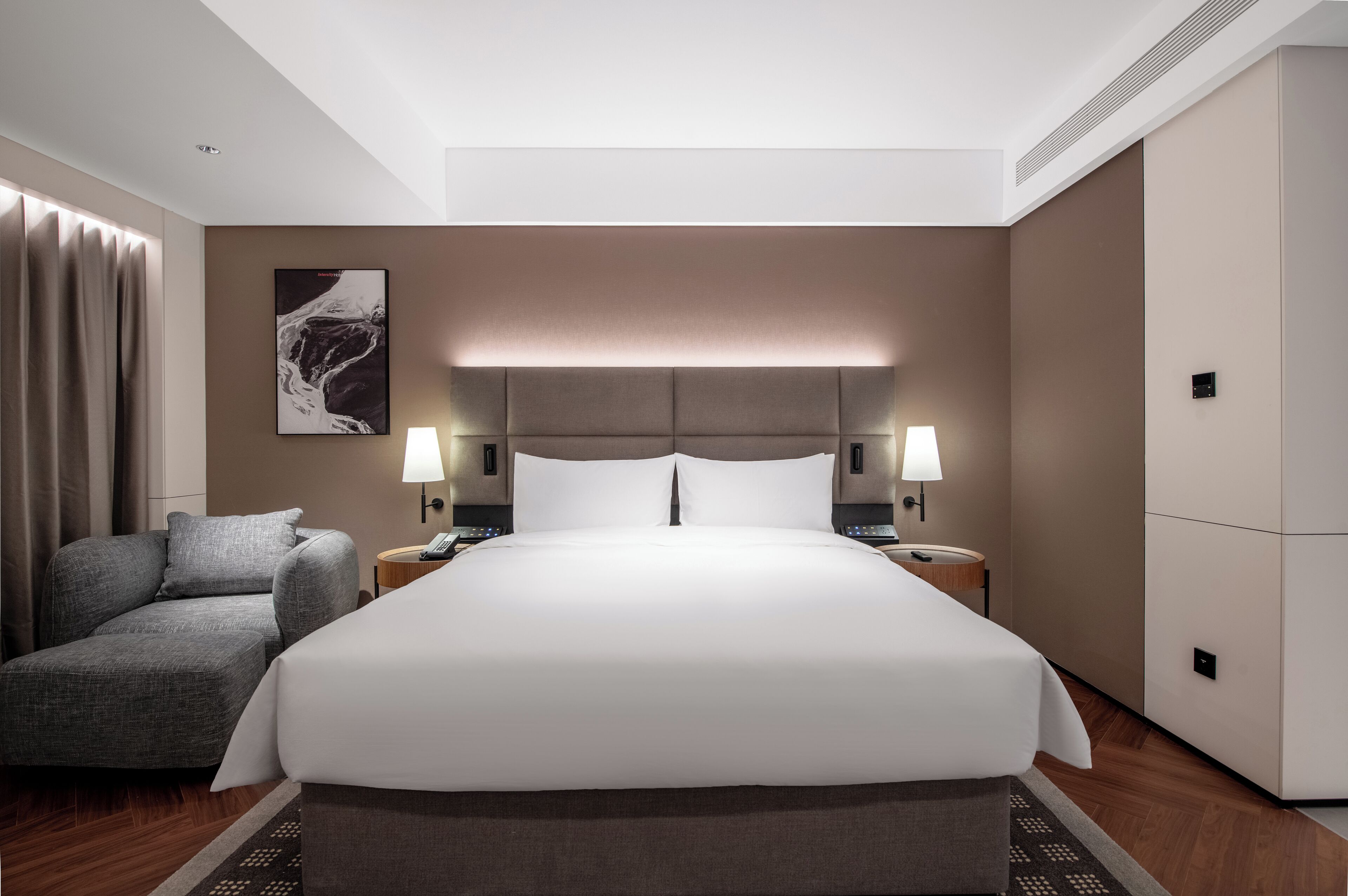 Quarto de hotel moderno com uma cama grande, duas mesas de cabeceira com luminárias, um sofá cinza, uma obra de arte na parede e piso de madeira.