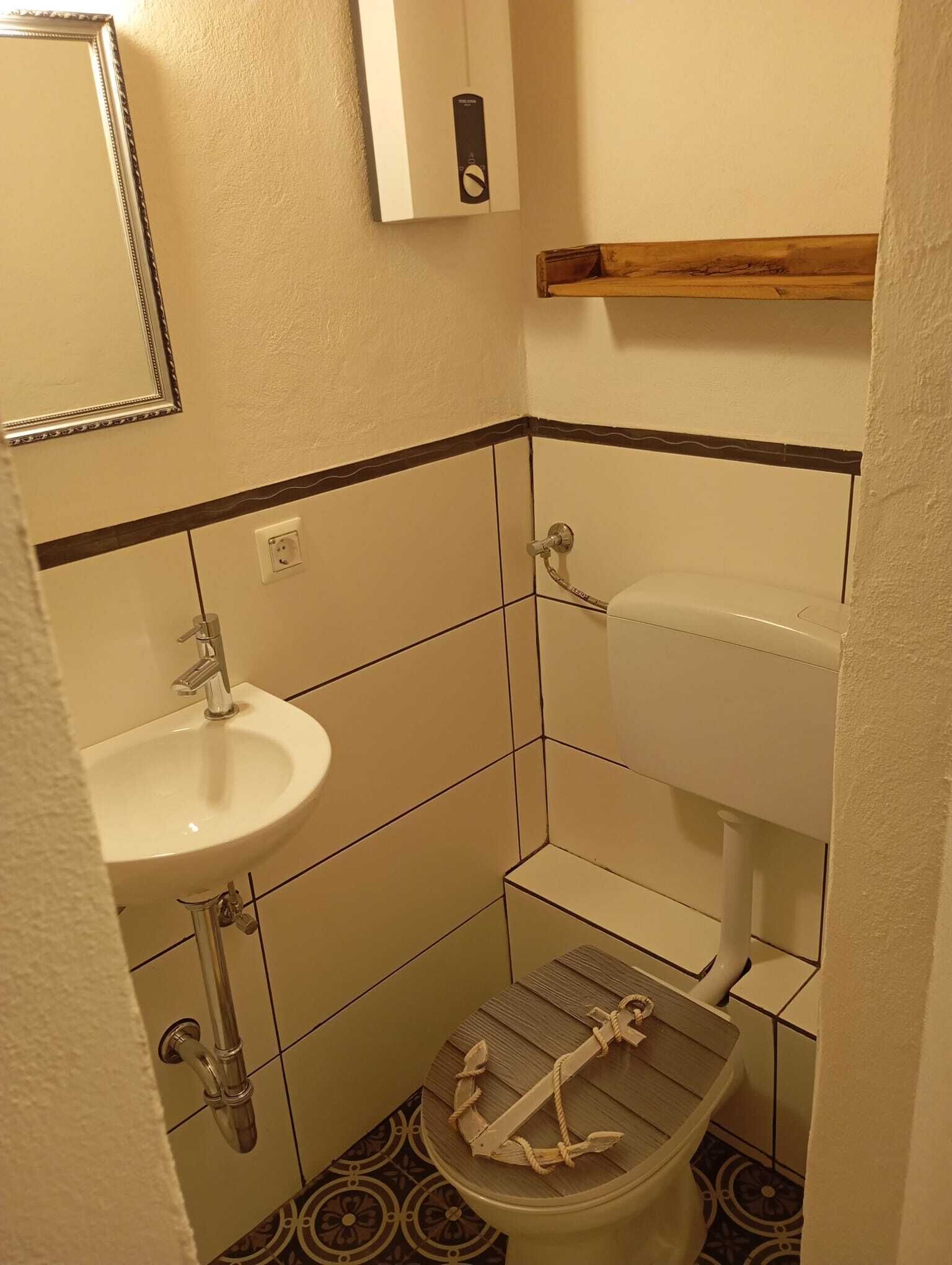 A small bathroom with a white sink, toilet, and a wooden shelf above the toilet.