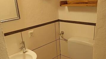 A small bathroom with a white sink, toilet, and a wooden shelf above the toilet.