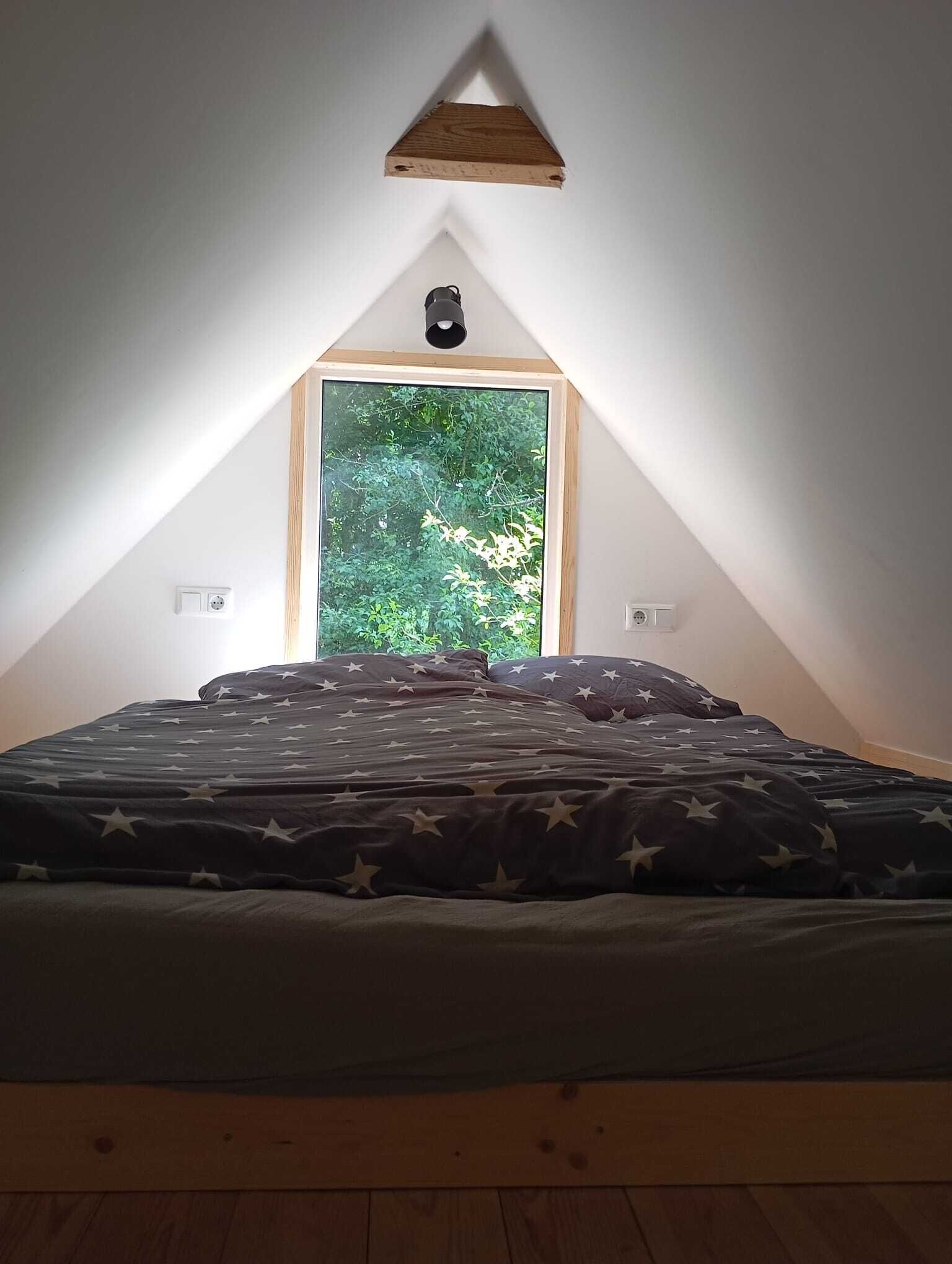 A bedroom with a sloped ceiling, a window with a view of greenery, a bed with a star-patterned bedspread, and a triangular wooden shelf above the window.