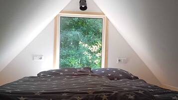 A bedroom with a sloped ceiling, a window with a view of greenery, a bed with a star-patterned bedspread, and a triangular wooden shelf above the window.