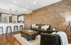 Ein modernes Wohnzimmer mit Backsteinwand, einer Couch, einem Couchtisch und einer Küchenzeile mit Hockern.