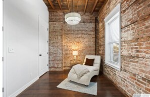 Ein Zimmer mit freiliegenden Backsteinwänden, einer weißen Tür, einem Bett mit Überwurfdecke, einer Lampe auf einem Nachttisch und einem Fenster mit Jalousien.