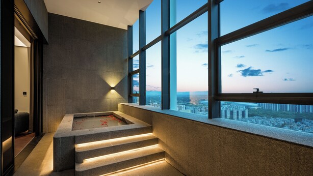 Un interior moderno con una bañera de piedra, vistas de la ciudad a través de amplios ventanales y una escalera que conduce a un nivel superior.