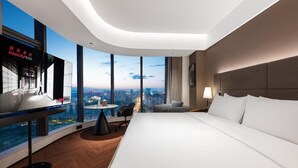 Una habitación de hotel moderna con una cama grande, un escritorio con una silla, una mesita pequeña y un televisor. La habitación tiene ventanas amplias que ofrecen vistas a la ciudad.