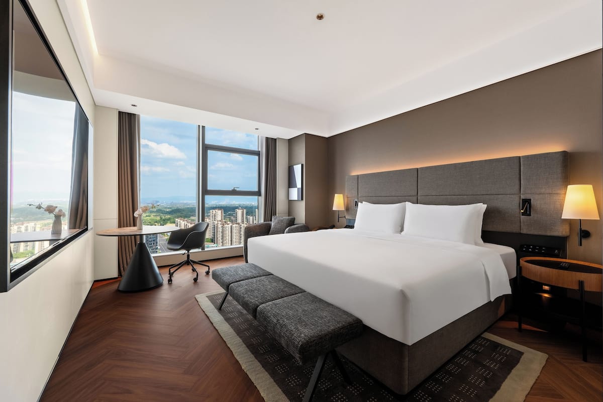 A modern hotel room with a large bed, a desk, a chair, and a view of the city.