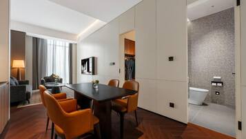A modern hotel room with a dining area, a sofa, a flat-screen TV, and a bathroom with a shower and toilet.