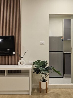 Ein modernes Wohnzimmer mit Flachbildfernseher, einer weißen Ablagetisch, einem Topf mit Pflanze und einer integrierten Küche mit Kühlschrank.
