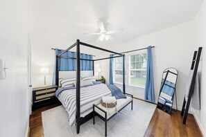 Ein Bett mit Baldachin, ein Deckenventilator, ein Nachttisch mit Lampe und ein Fenster mit Vorhängen.