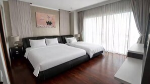 Quarto de hotel com duas camas, uma escrivaninha, uma lâmpada, um quadro e uma janela grande com cortinas.