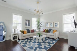 Ein Wohnzimmer mit einem weißen Sofa, bunten Kissen, einem blau-weißen Teppich, einer hohen Pflanze und gerahmten Bildern an der Wand.