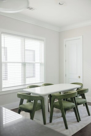 Ein Zimmer mit einem weißen Tisch, grünen Stühlen, einem Fenster mit Jalousien und einer weißen Tür.
