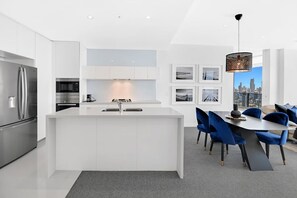 A modern kitchen with a central island, stainless steel appliances, and a dining area with blue chairs and a city view. - Ocean Luxury at Oracle Level 32 (Gold Coast City)