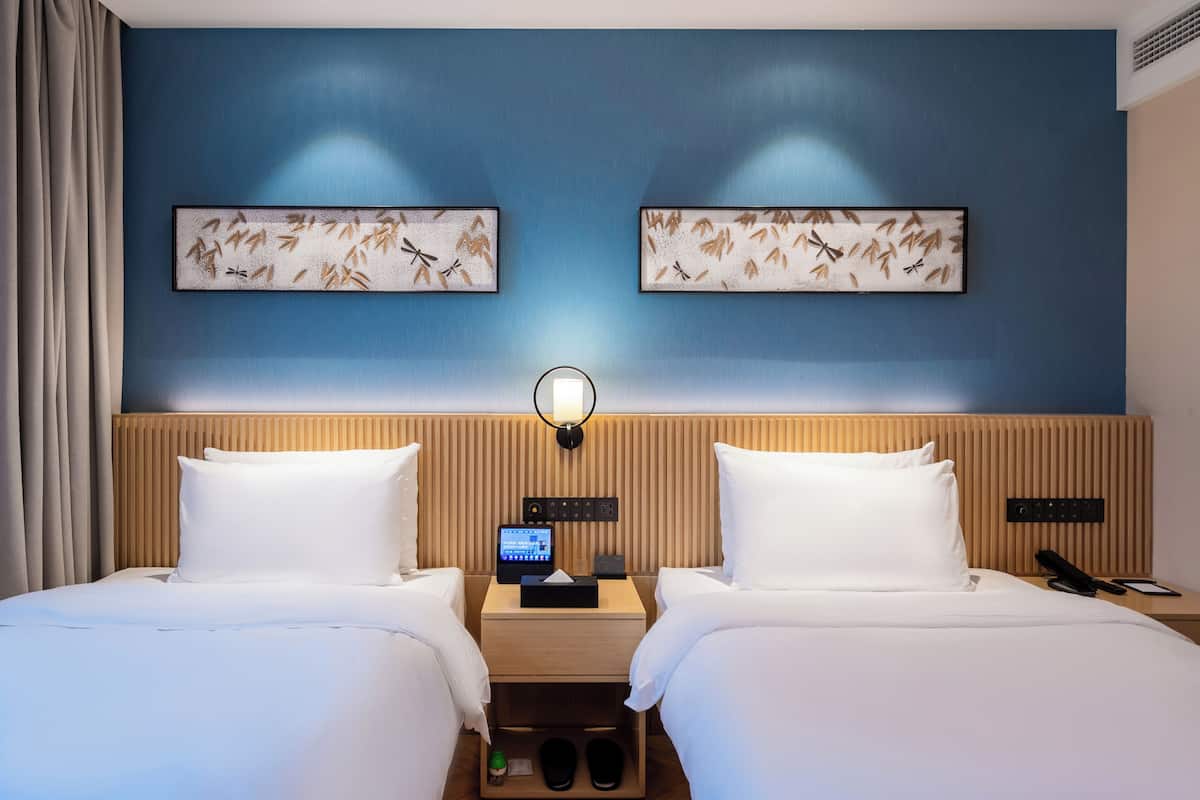 Una habitación de hotel con dos camas, cabecera de madera, mesita de noche con lámpara, reloj digital y una obra de arte en la pared.
