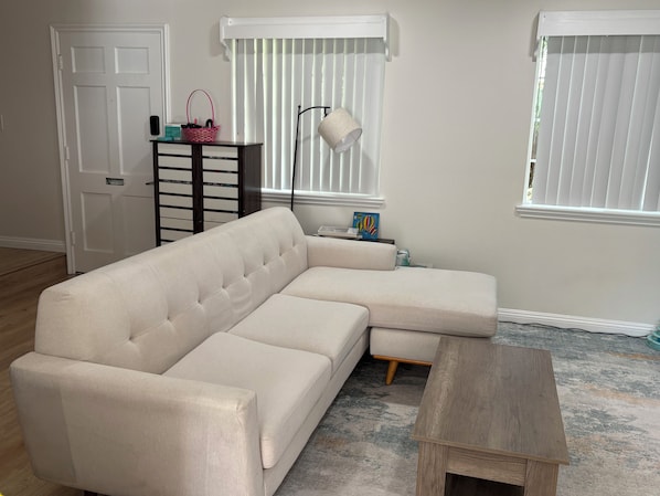 Ein Wohnzimmer mit Ecksofa, einem kleinen Holztisch, einer Stehlampe, einem Korb und einer Heizung.
