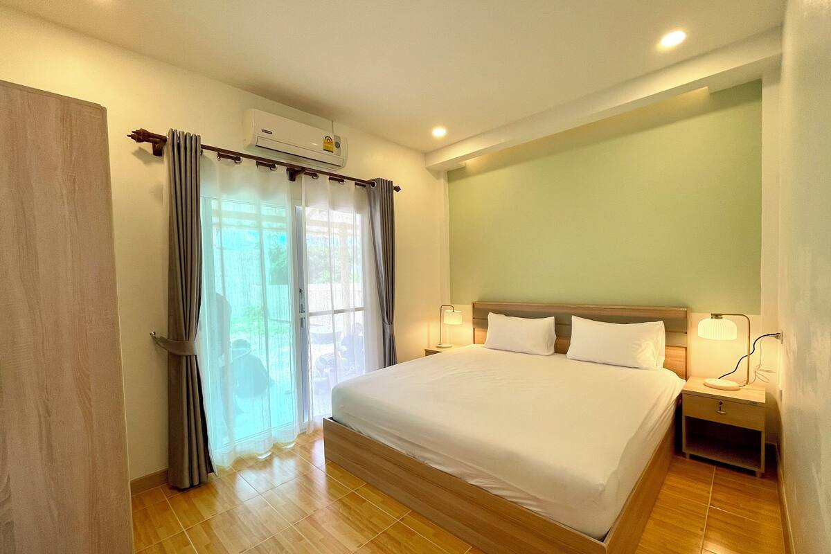 Habitación de hotel con una cama grande, una mesita de noche con lámpara, un equipo de aire acondicionado y vistas al exterior a través de una ventana con cortinas.