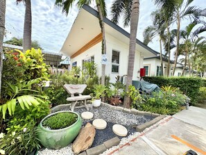 Un edificio moderno de fachada blanca, rodeado de plantas tropicales y un pequeño jardín con una silla decorativa y una fuente de agua.