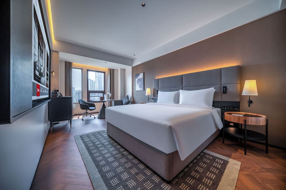 A modern hotel room with a large bed, a desk, a chair, and a view of the city.
