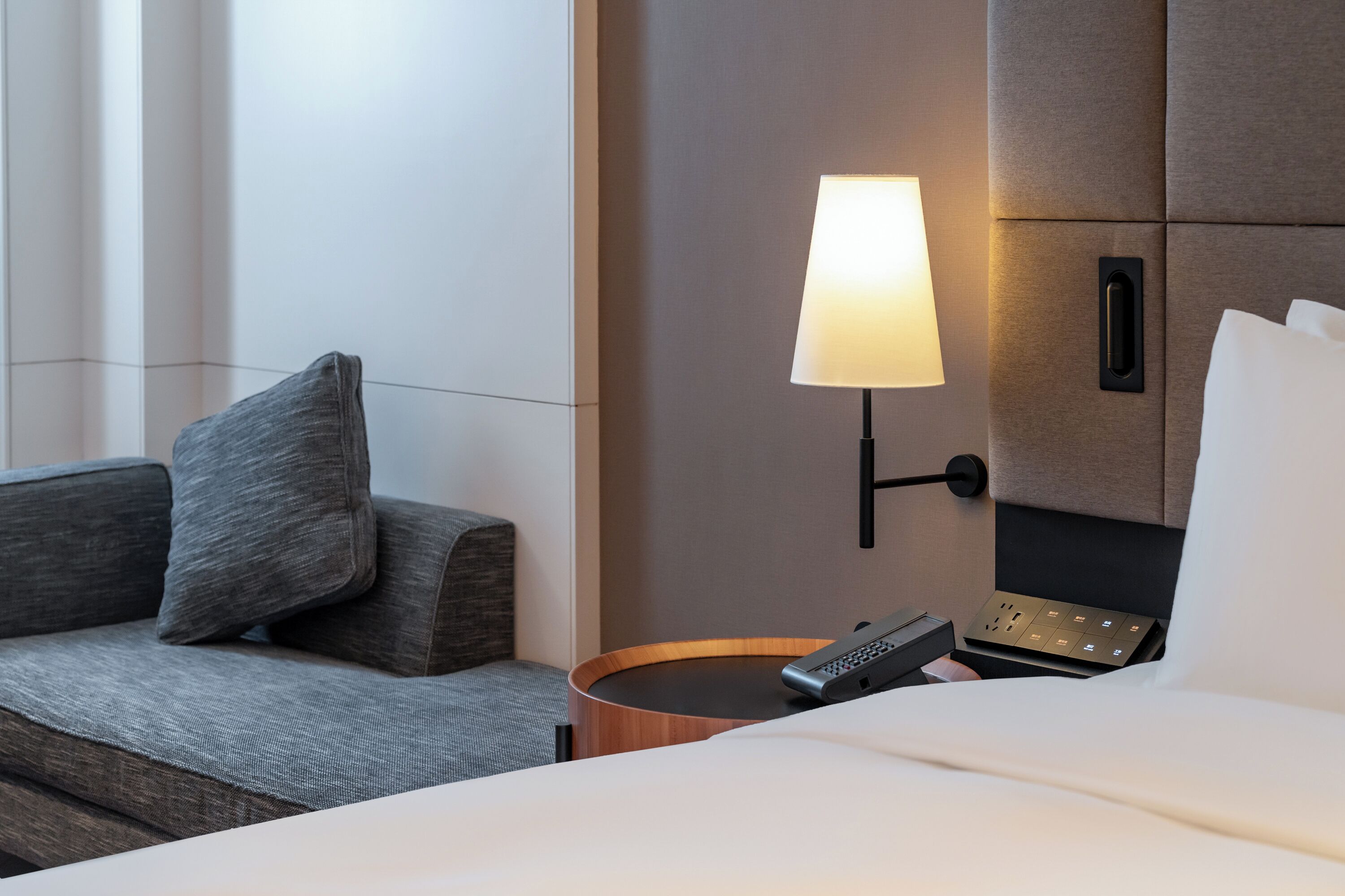 A hotel room with a bed, a bedside table with a lamp, a remote control, and a wall-mounted control panel.
