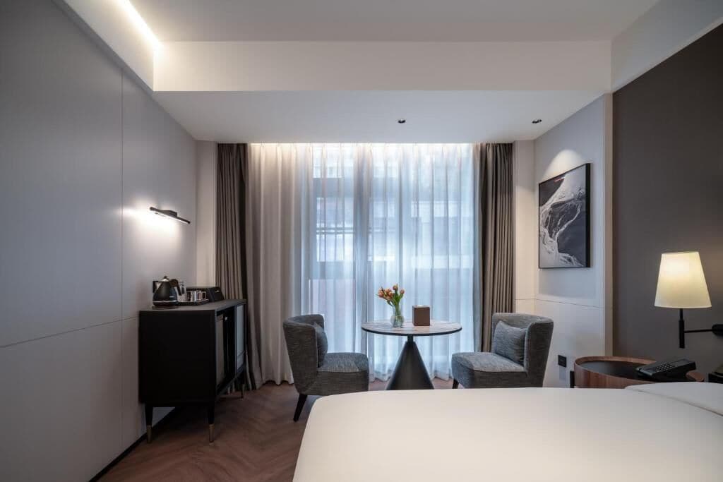 Habitación de hotel con cama, escritorio, dos sillas, una mesita con flores, un cuadro en la pared y una lámpara.