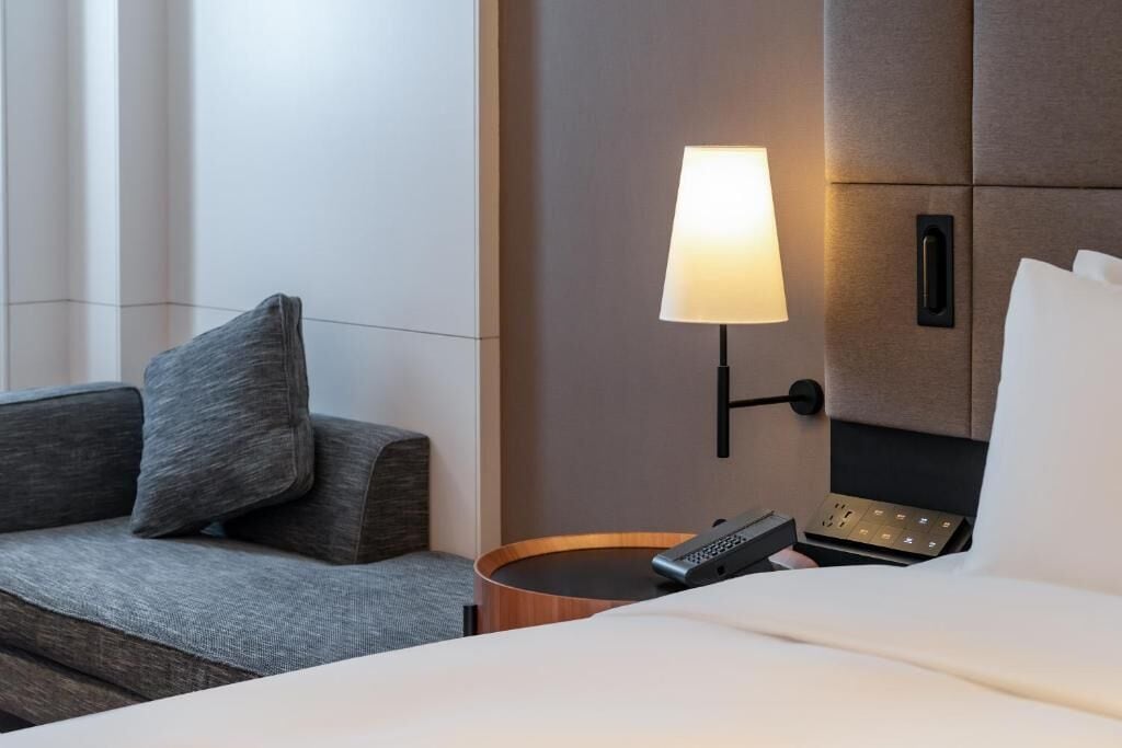 Ein Hotelzimmer mit Bett, Nachttisch mit Lampe, Fernbedienung und einem kleinen Holztisch.