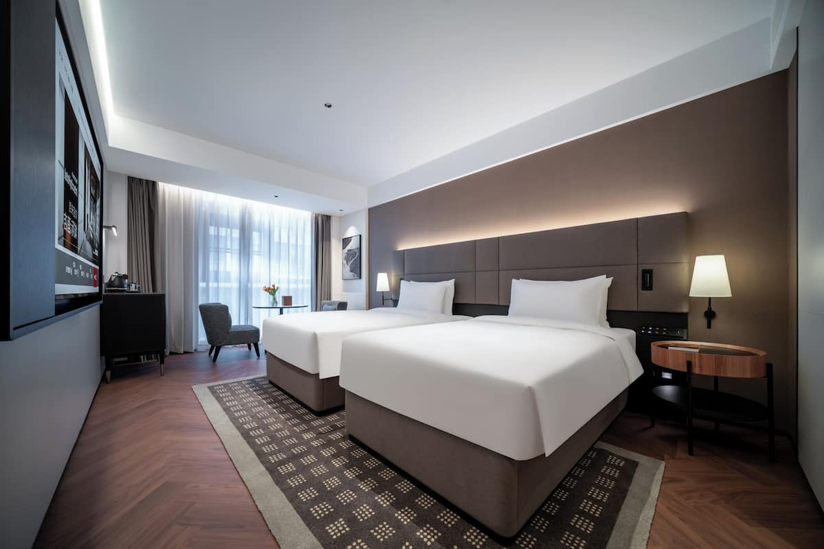 A hotel room with two beds, a desk, a chair, a TV, and a window with curtains.