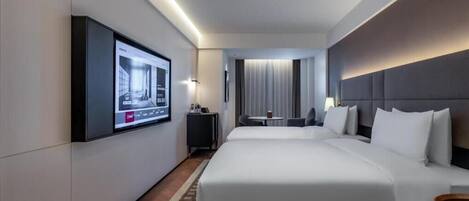 A hotel room with a large bed, a TV mounted on the wall, a small table with a chair, and a view of the city through the window.