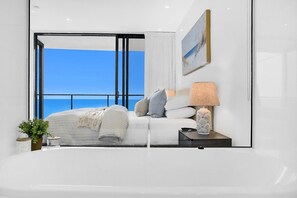 A modern bedroom with a large window, a bed, a bedside table with a lamp, and a small plant. - Ocean Front Luxe at Oracle Level 34 (Gold Coast City)
