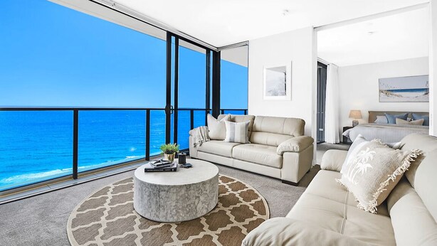 Ein modernes Wohnzimmer mit großem Fenster zum Meer, einem runden Couchtisch und einem beigefarbenen Sofa.