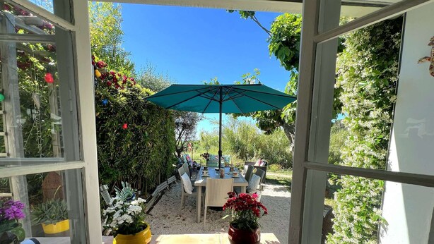 Ventana abierta con vistas a un patio, que incluye una mesa y sillas, un parasol verde y plantas en macetas.
