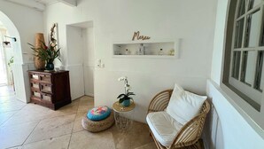 Una habitación con una silla de mimbre, una pequeña mesa redonda, una planta en maceta, un estante decorativo con un cartel y una puerta que da a otra habitación.