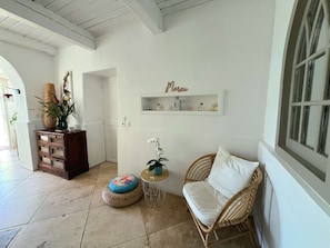 Une pièce comprenant une chaise en osier, une petite table ronde, une plante en pot, une étagère décorative avec une inscription, et une porte donnant sur une autre pièce.