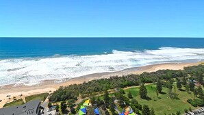 Une vue côtière comprenant une plage, des vagues de l’océan et un espace vert avec des arbres et un bâtiment.