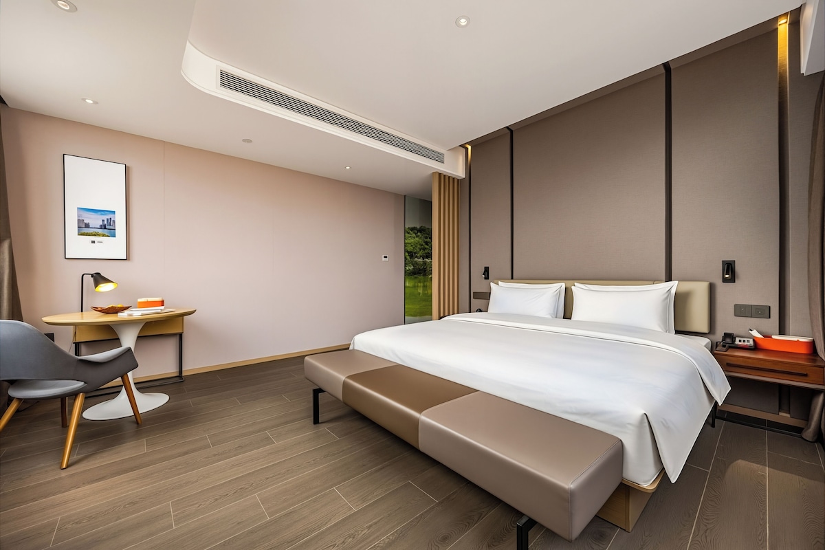 A modern hotel room with a bed, bedside table, desk, chair, and a view of greenery outside.