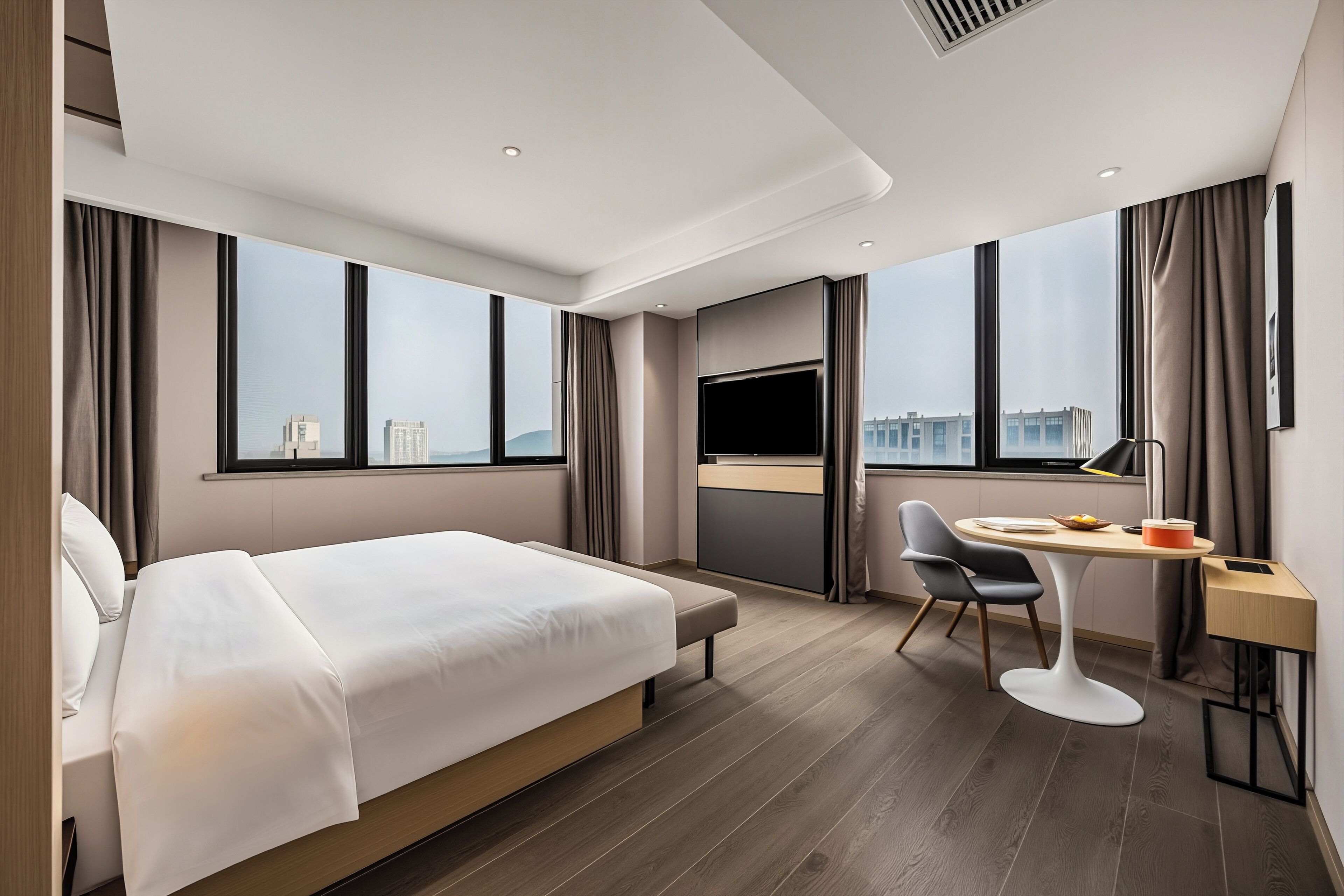 A modern hotel room with a large bed, a desk with a chair, a television, and a view of the city.