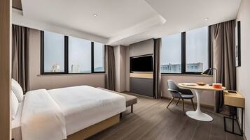 A modern hotel room with a large bed, a desk with a chair, a television, and a view of the city.
