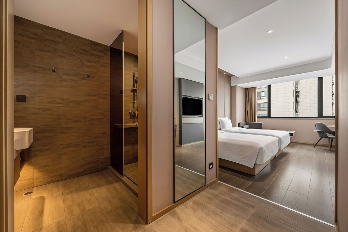 A modern hotel room with a wooden floor, a large mirror, a bed with two pillows, a TV, and a window with curtains.