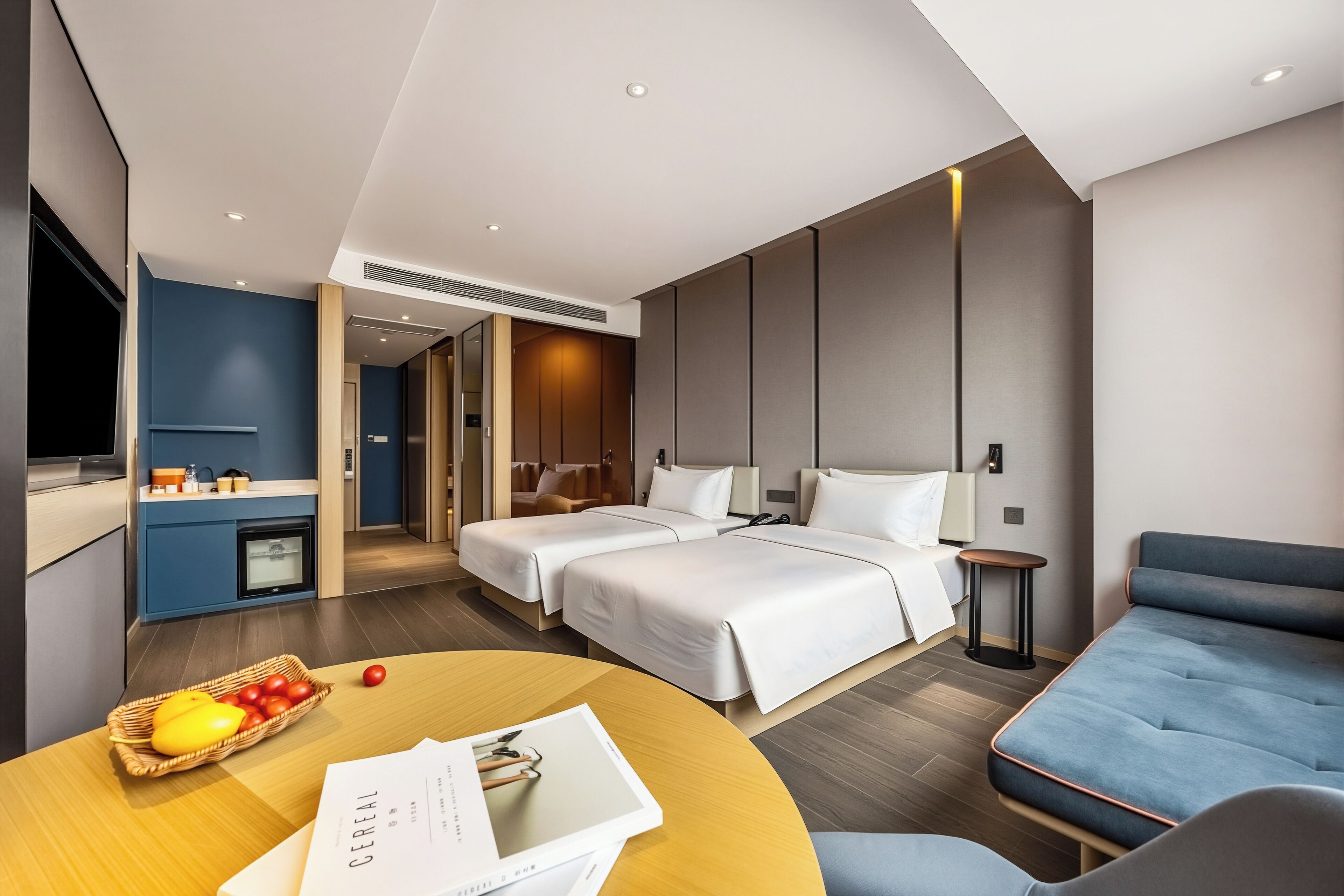 A modern hotel room with two beds, a dining table with fruit, a TV, and a kitchenette.
