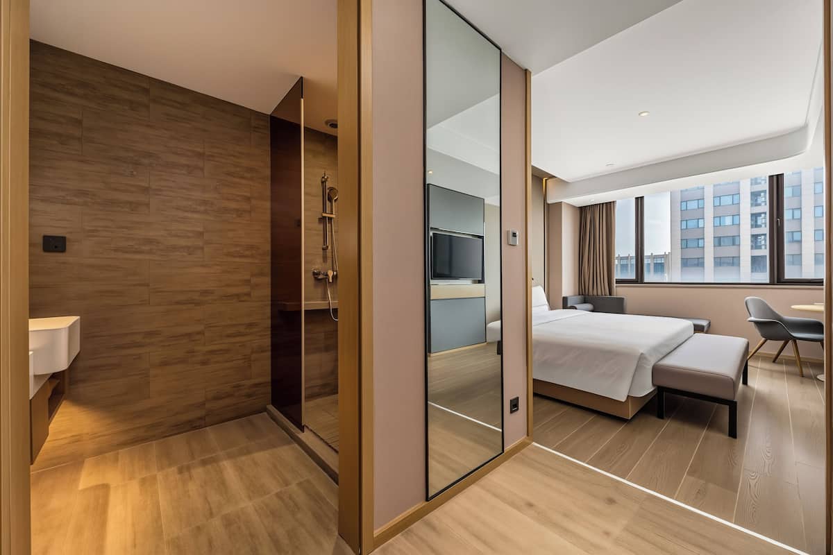 A modern hotel room with a wooden floor, a bed with a bench, a desk, a chair, and a large window with a city view.