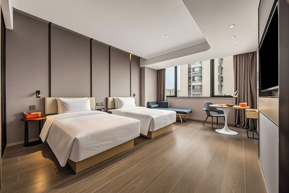A modern hotel room with two beds, a desk, a chair, a TV, and a window with a city view.