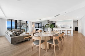 Ein modernes Wohnzimmer mit Sofa, Esstisch, Stühlen, einer Küchenzeile und großen Fenstern mit Aussicht.