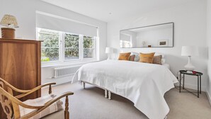 Un dormitorio con un ventanal grande, una cama, una silla de madera, una mesita de noche y un espejo.