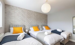 A hotel room with three beds, each with a grey headboard, white bedding, and a yellow pillow. There is a white radiator, a bedside table with a lamp, and a hanging light fixture.
