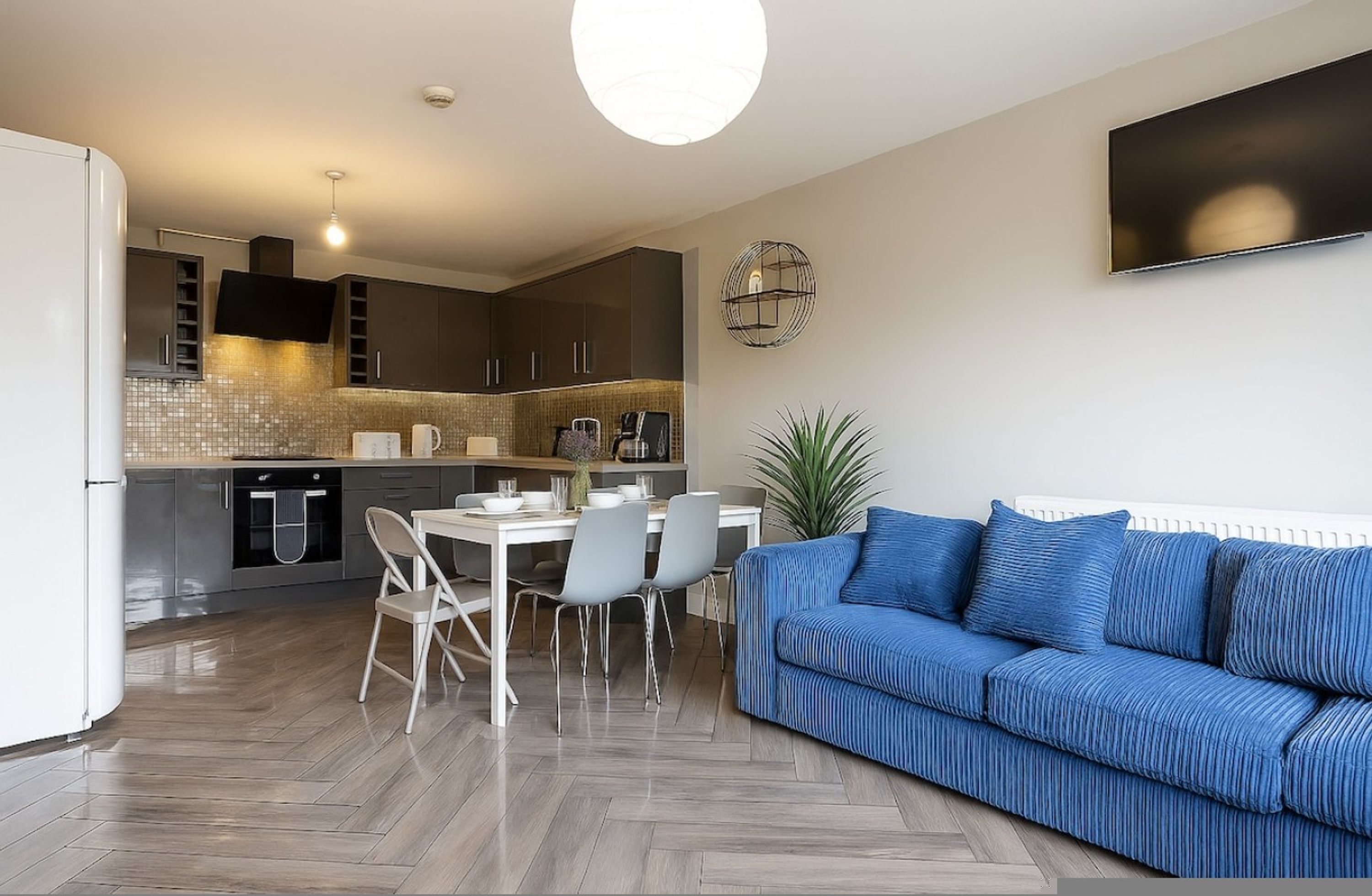 A modern kitchen with a dining area, a blue sofa, and a large TV on the wall.