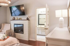 A bedroom with a TV, fireplace, bed, and a bathroom with a mirror and sink.