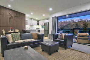 Una sala moderna con sofá, sillones y una mesa de centro. Se ve una cocina al fondo y un ventanal con vistas a la montaña.